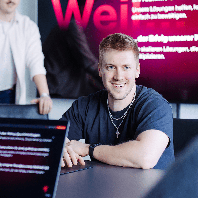 Eine Person sitzt l&auml;chelnd an einem Konferenztisch mit Laptop, im Hintergrund l&auml;uft eine Pr&auml;sentation auf einem gro&szlig;en Bildschirm.