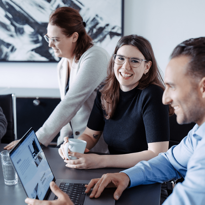 Drei Personen sitzen gemeinsam an einem Tisch im Besprechungsraum, l&auml;cheln und arbeiten an Laptops.